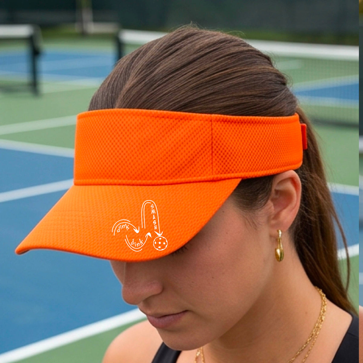 A close-up lifestyle shot of a bright orange athletic visor with a white pickleball 'DINK DINK SMASH' logo with a pickleball graphic