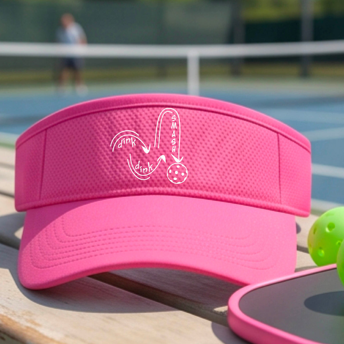 A close-up lifestyle shot of a bright pink athletic visor with a white pickleball 'DINK DINK SMASH' logo with a pickleball graphic