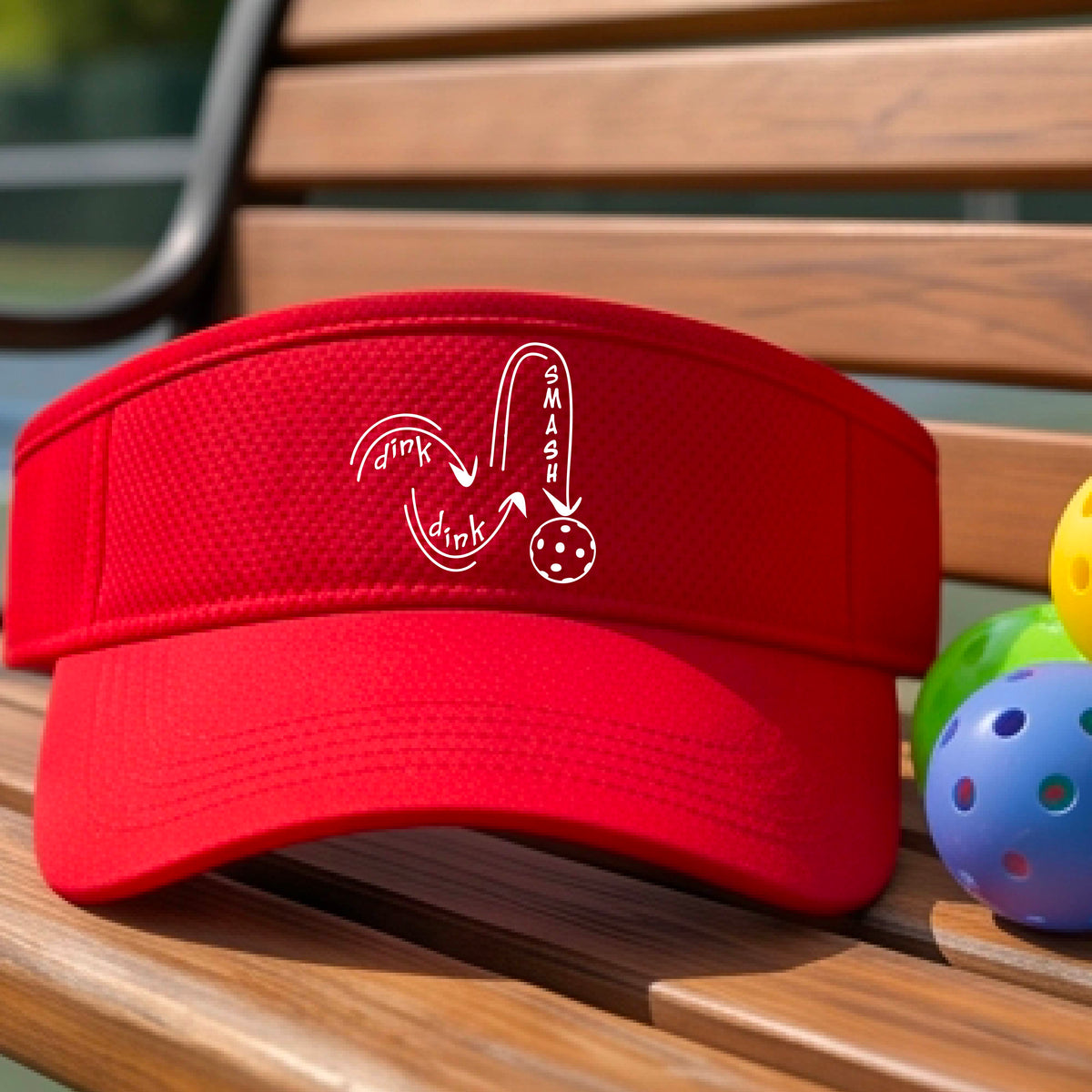A close-up lifestyle shot of a bright red athletic visor with a white pickleball 'DINK DINK SMASH' logo with a pickleball graphic
