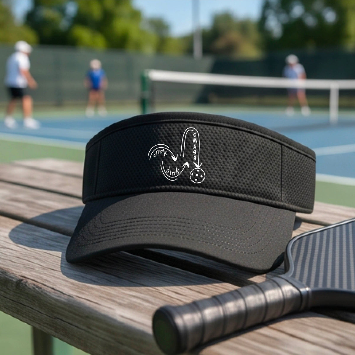 A close-up lifestyle shot of a black athletic visor with a white pickleball 'DINK DINK SMASH' logo with a pickleball graphic