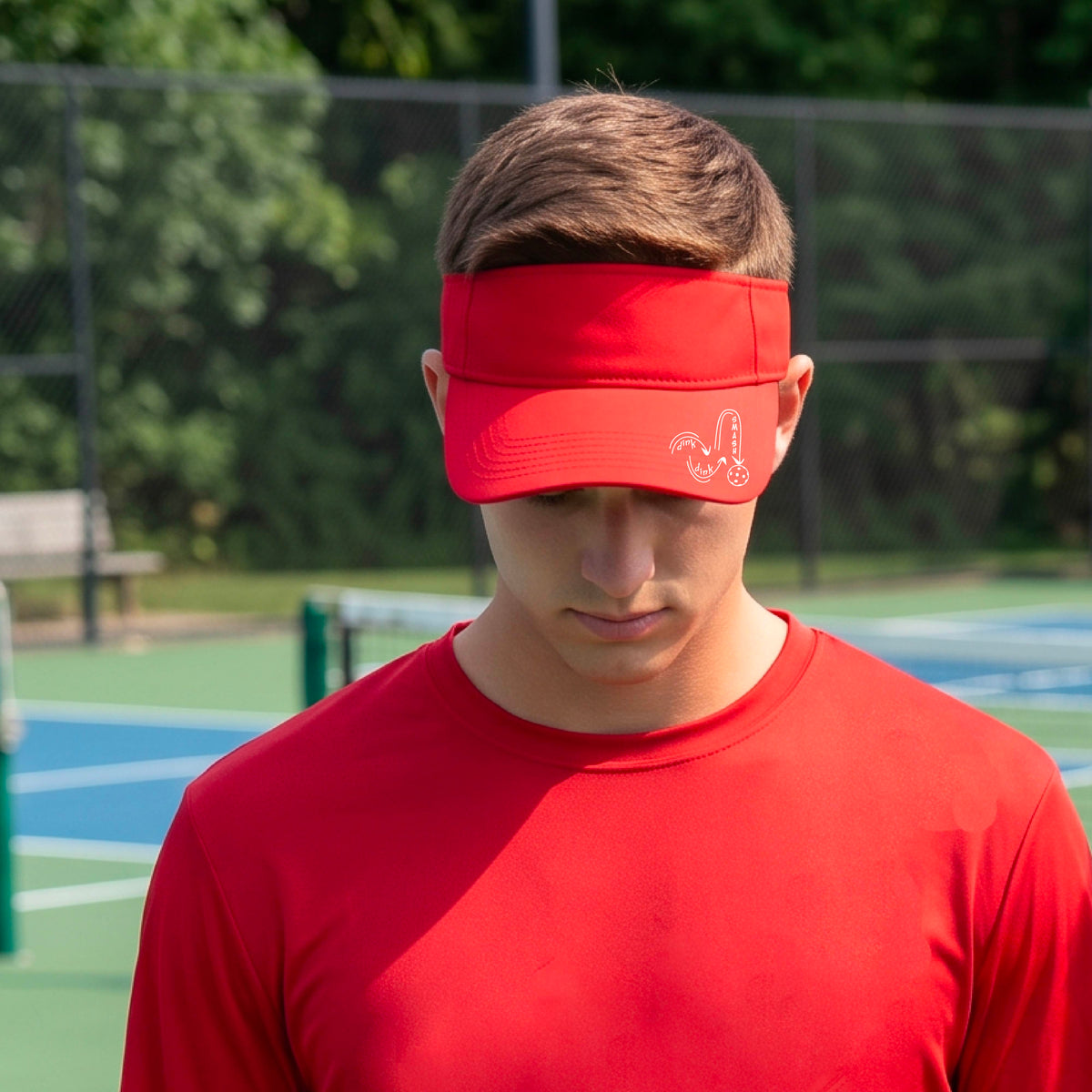 A close-up lifestyle shot of a bright red athletic visor with a white pickleball 'DINK DINK SMASH' logo with a pickleball graphic
