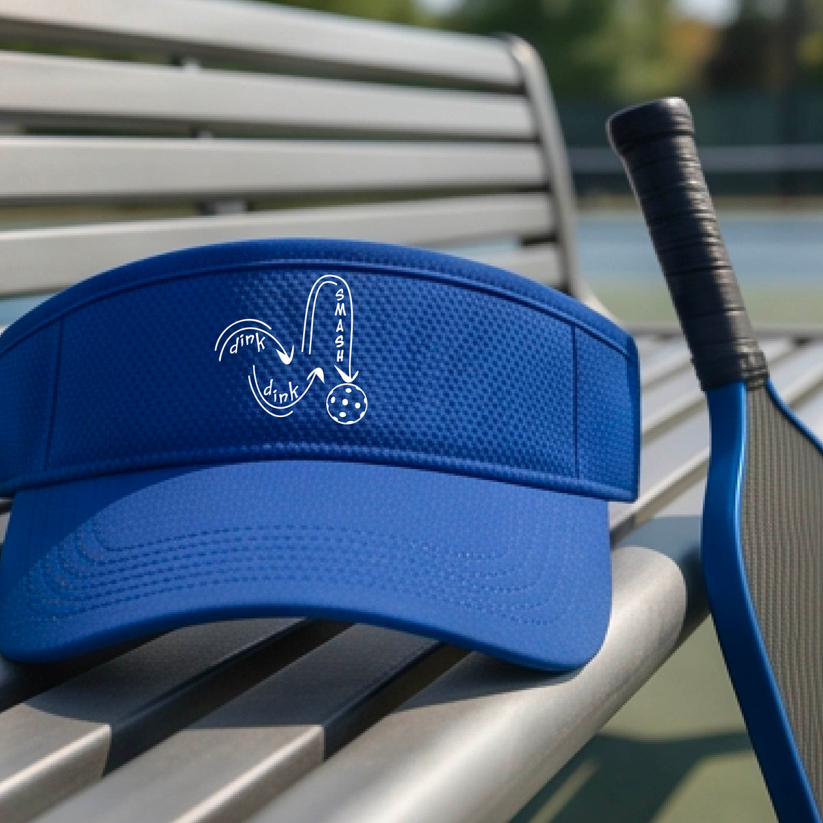A close-up lifestyle shot of a bright blue athletic visor with a white pickleball 'DINK DINK SMASH' logo with a pickleball graphic