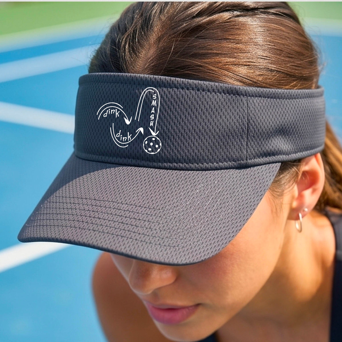 A close-up lifestyle shot of a gray athletic visor with a white pickleball 'DINK DINK SMASH' logo with a pickleball graphic