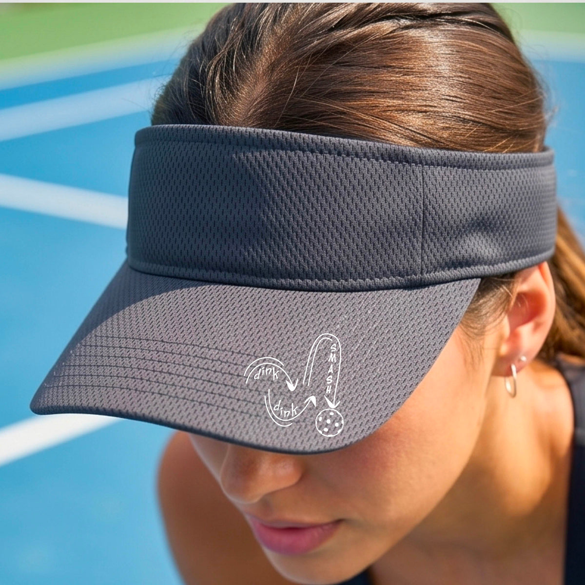 A close-up lifestyle shot of a gray athletic visor with a white pickleball 'DINK DINK SMASH' logo with a pickleball graphic