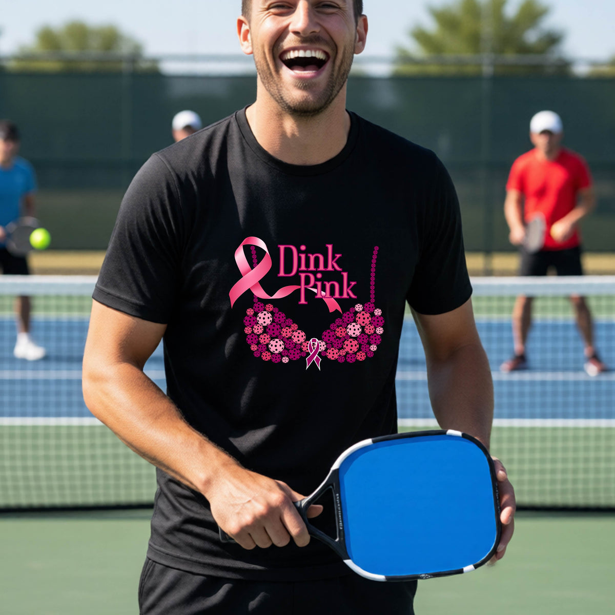 A man on a pickleball court wearing a black t-shirt with a pink 'Dink Pink' bra graphic and floral design, with other players blurred in the background.