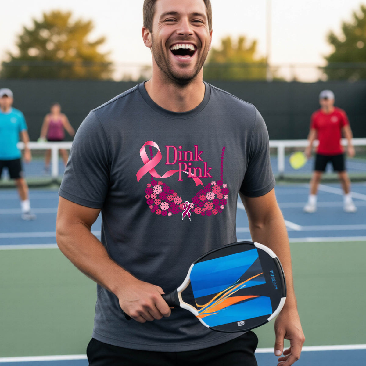 A man on a pickleball court wearing a gray  t-shirt with a pink 'Dink Pink' bra graphic and floral design, with other players blurred in the background.