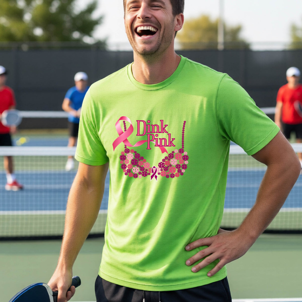 A man on a pickleball court wearing a bright lime green t-shirt with a pink 'Dink Pink' bra graphic and floral design, with other players blurred in the background.