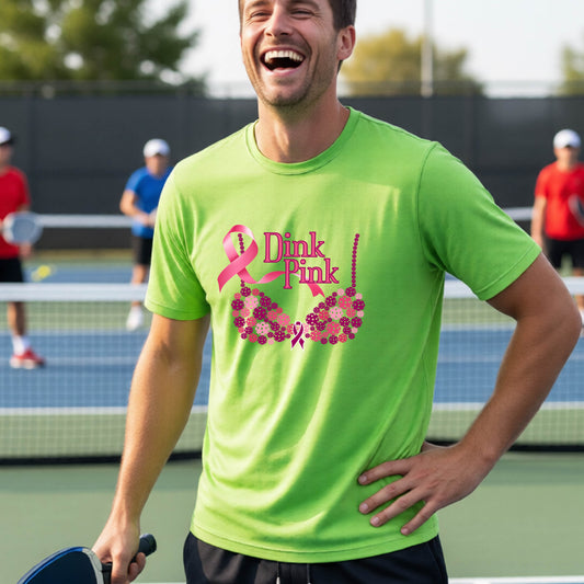 A man on a pickleball court wearing a bright lime green t-shirt with a pink 'Dink Pink' bra graphic and floral design, with other players blurred in the background.