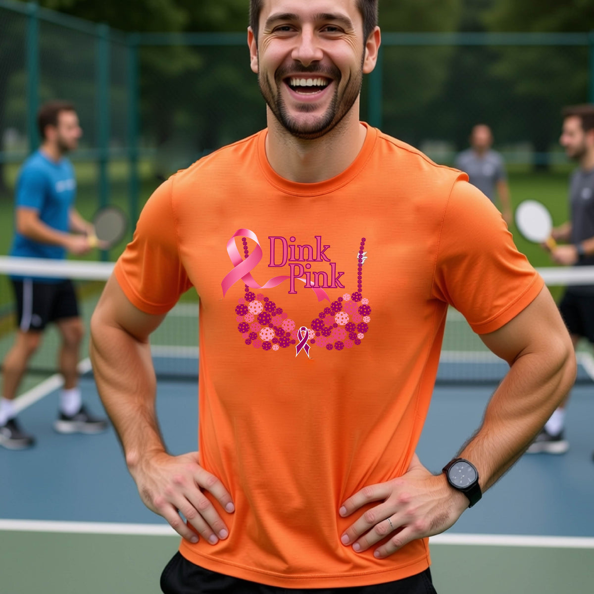 A man on a pickleball court wearing ann orange t-shirt with a pink 'Dink Pink' bra graphic and floral design, with other players blurred in the background.