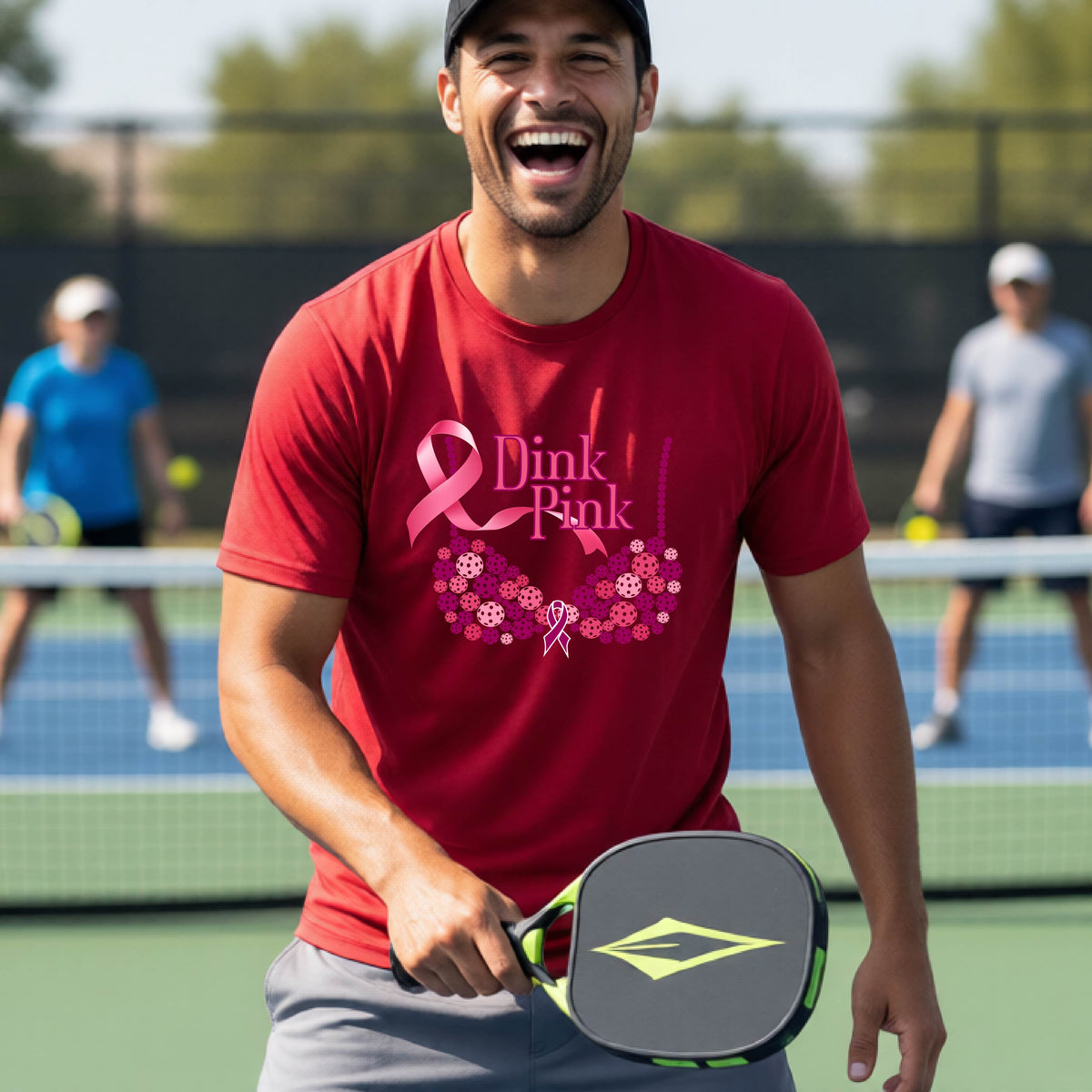 A man on a pickleball court wearing a red t-shirt with a pink 'Dink Pink' bra graphic and floral design, with other players blurred in the background.
