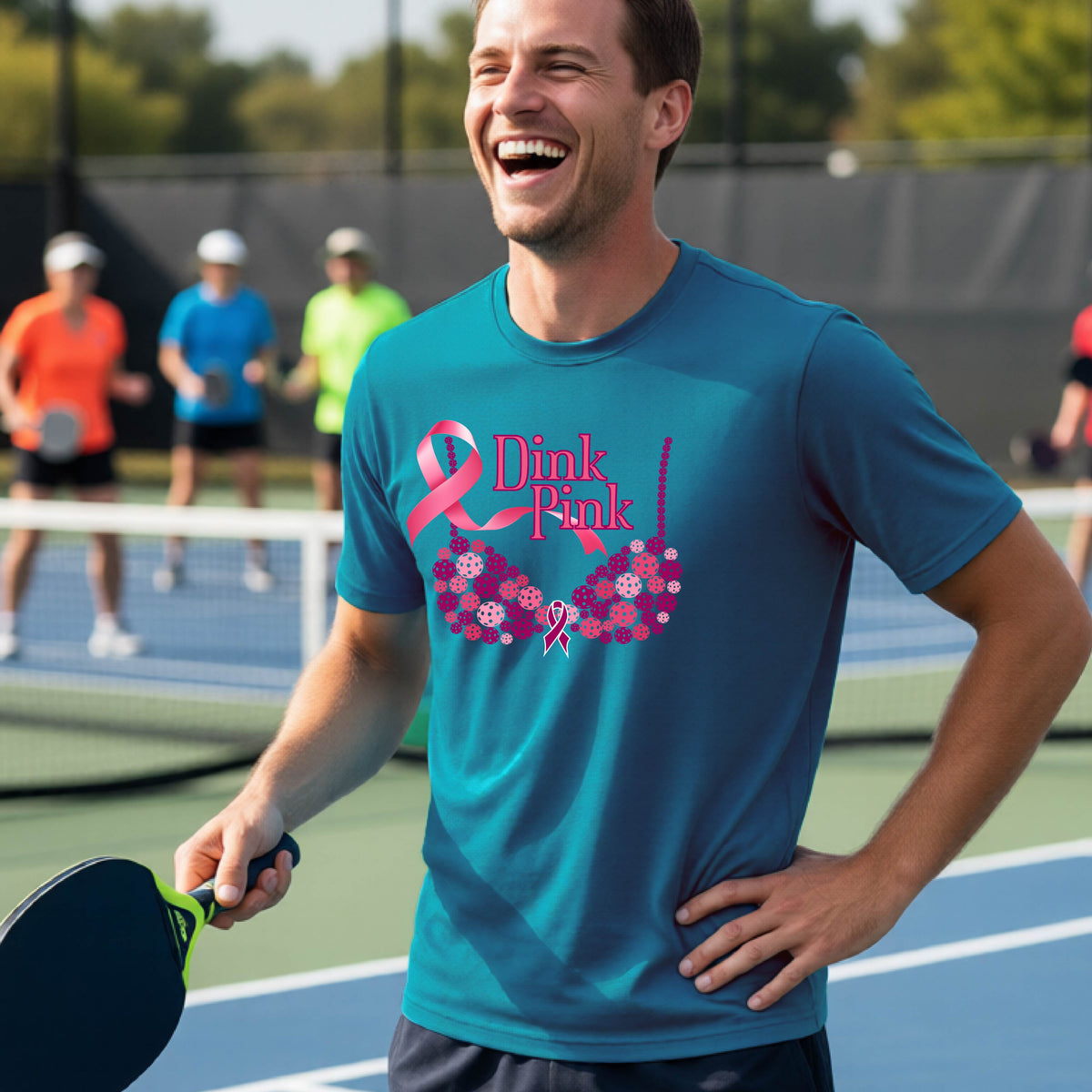 A man on a pickleball court wearing a teal t-shirt with a pink 'Dink Pink' bra graphic and floral design, with other players blurred in the background.