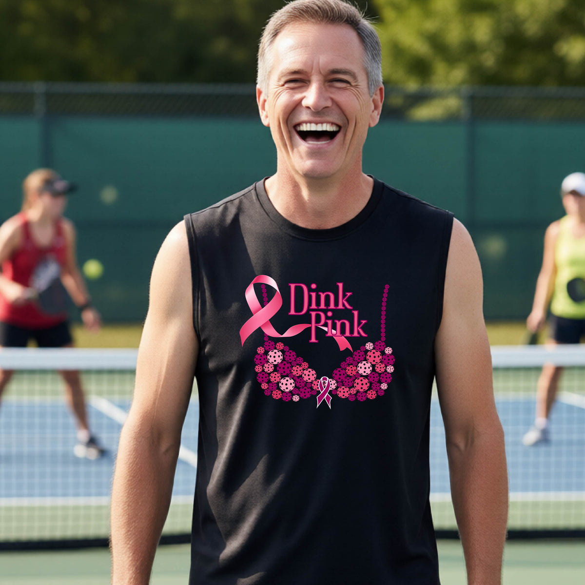 A smiling man wearing a black sleeveless tank top with a pink 'Dink Pink' breast cancer awareness graphic stands on an outdoor pickleball court. 