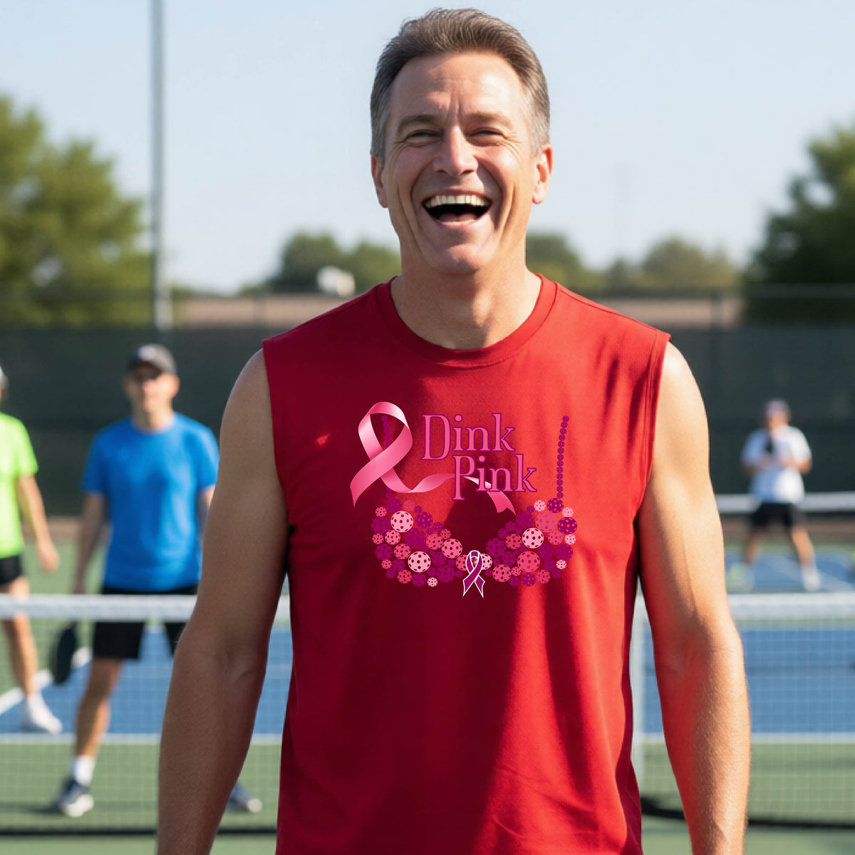A smiling man wearing a red sleeveless tank top with a pink 'Dink Pink' breast cancer awareness graphic stands on an outdoor pickleball court. 