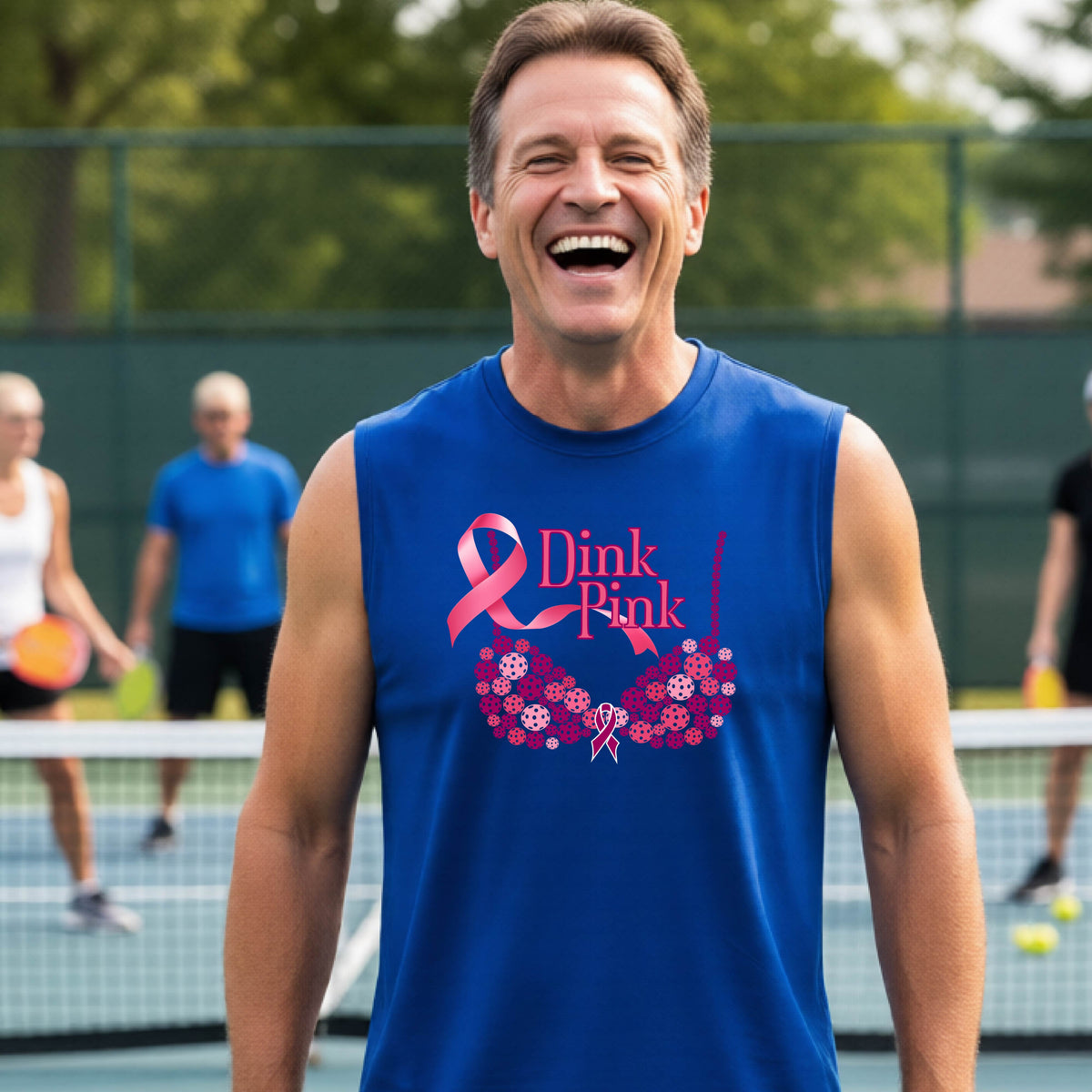 A smiling man wearing a blue sleeveless tank top with a pink 'Dink Pink' breast cancer awareness graphic stands on an outdoor pickleball court. 