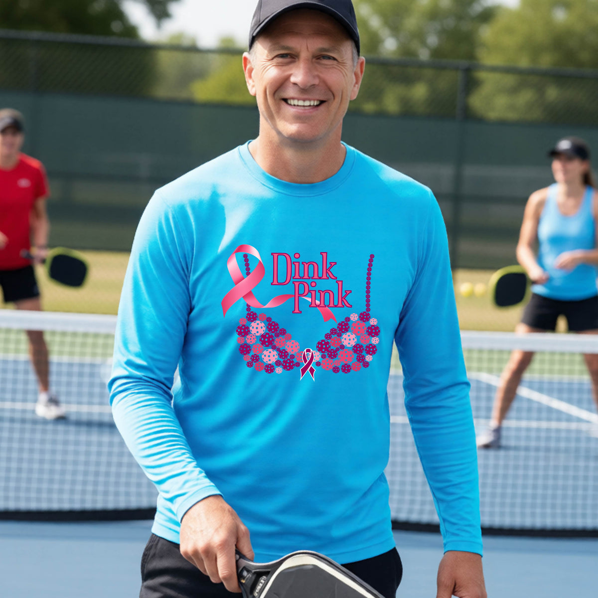 A smiling man wearing a bright blue long-sleeve shirt with a pink 'Dink Pink' breast cancer awareness graphic stands on a pickleball court.