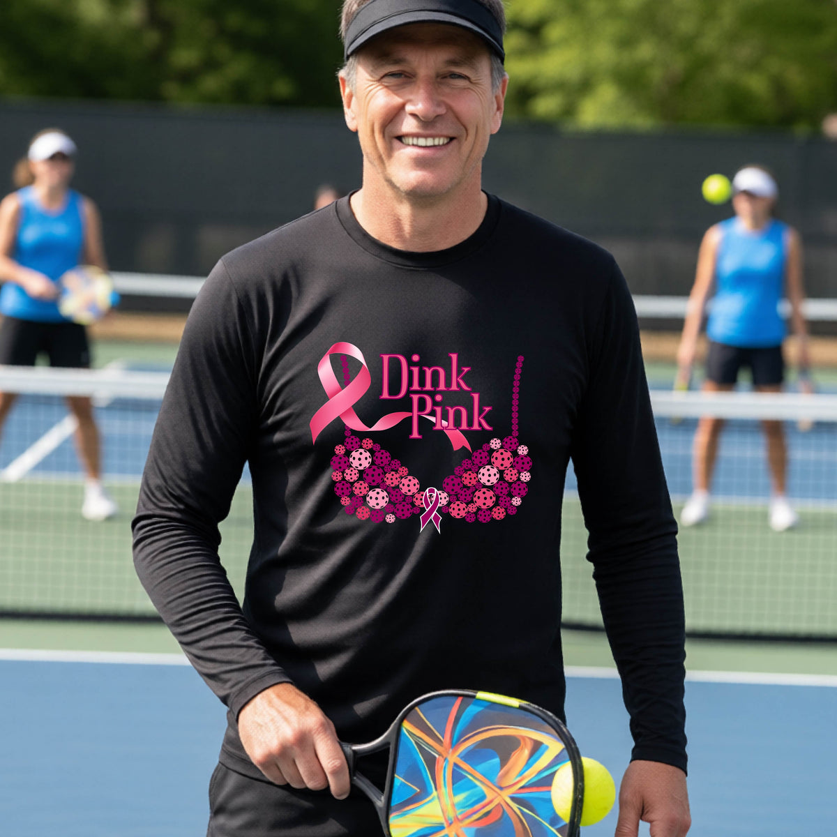 A smiling man wearing a black long-sleeve shirt with a pink 'Dink Pink' breast cancer awareness graphic stands on a pickleball court.
