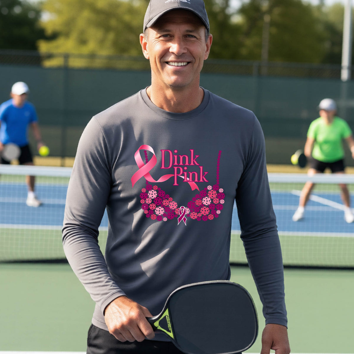 A smiling man wearing a gray long-sleeve shirt with a pink 'Dink Pink' breast cancer awareness graphic stands on a pickleball court.