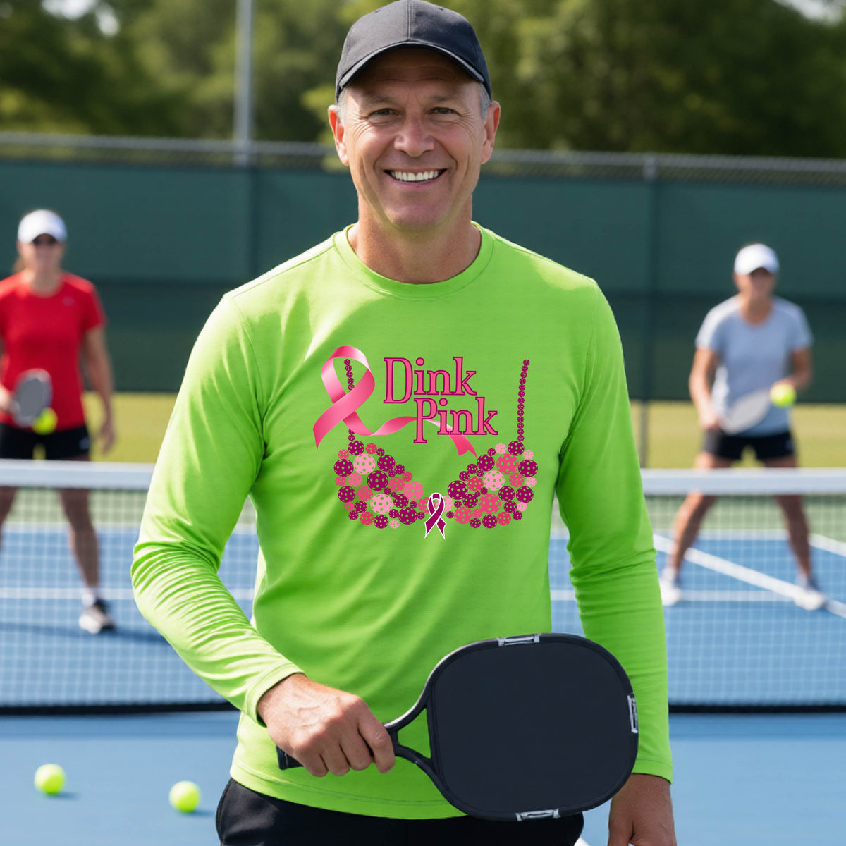 A smiling man wearing a lime green long-sleeve shirt with a pink 'Dink Pink' breast cancer awareness graphic stands on a pickleball court.