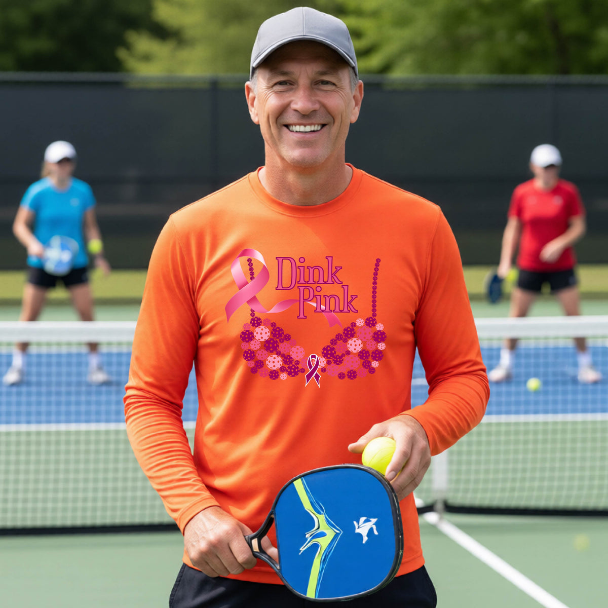 A smiling man wearing an orange long-sleeve shirt with a pink 'Dink Pink' breast cancer awareness graphic stands on a pickleball court.