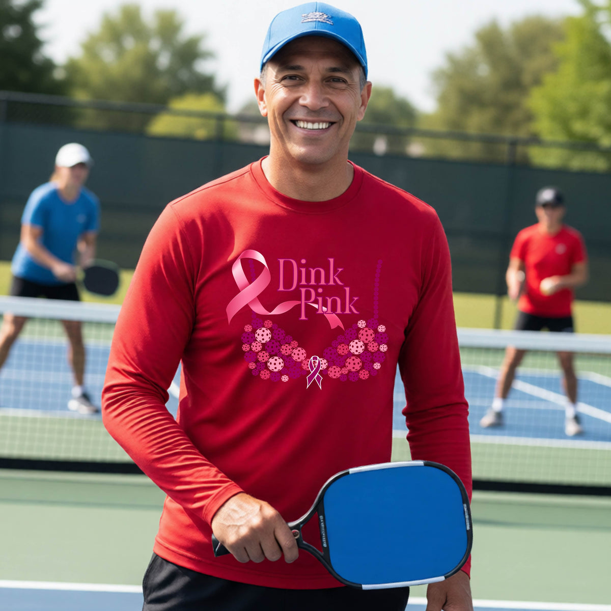 A smiling man wearing a red long-sleeve shirt with a pink 'Dink Pink' breast cancer awareness graphic stands on a pickleball court.