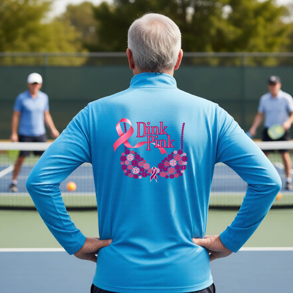 A man on a pickleball court seen from behind wearing a bright blue long-sleeve quarter-zip shirt with a large pink 'Dink Pink' breast cancer awareness graphic made of pickleballs on the back.