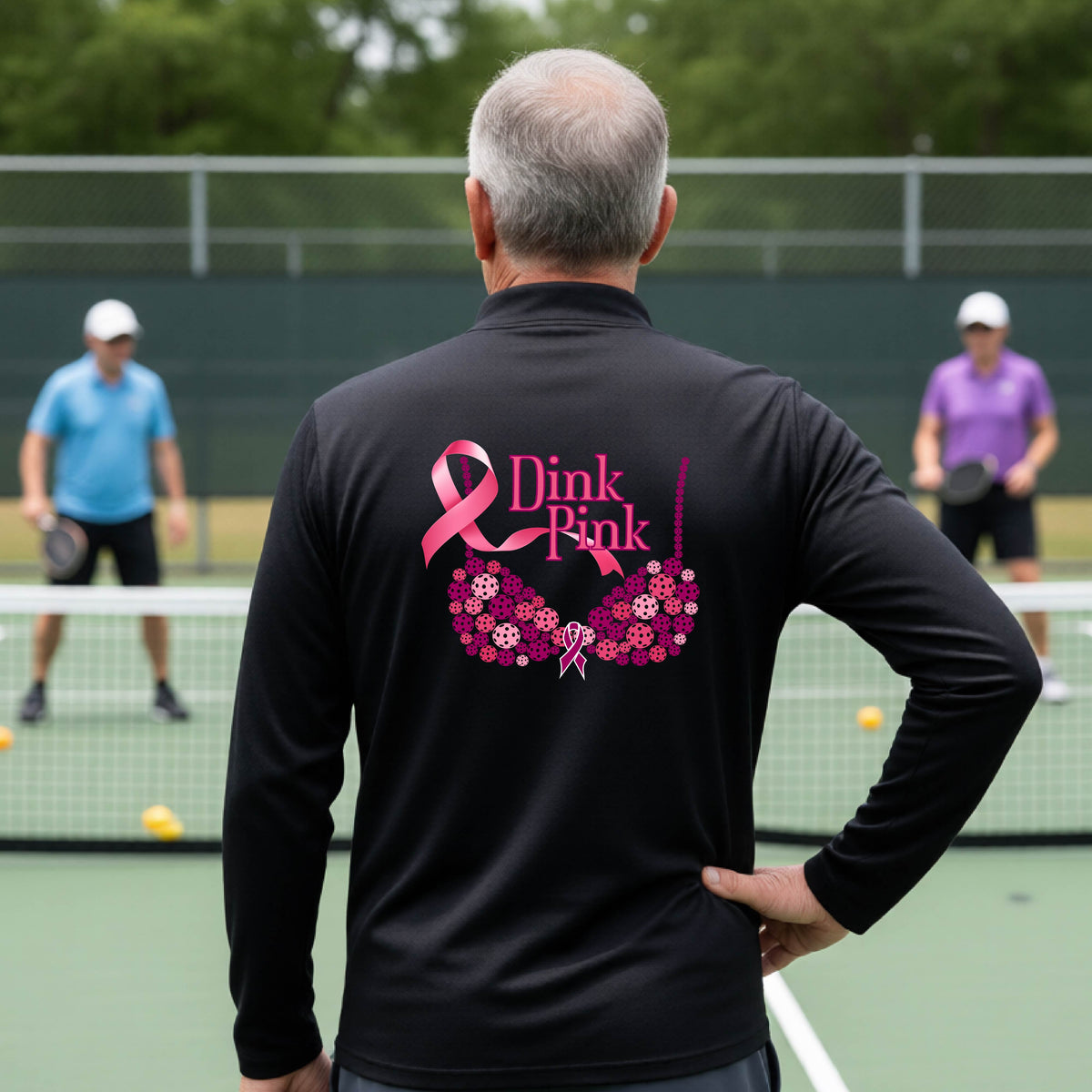 A man on a pickleball court seen from behind wearing a blacklong-sleeve quarter-zip shirt with a large pink 'Dink Pink' breast cancer awareness graphic made of pickleballs on the back.