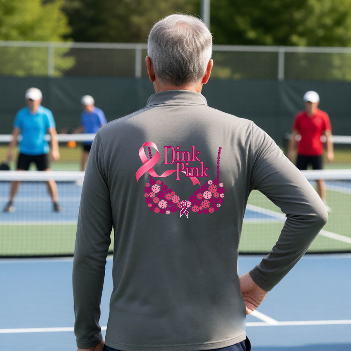 A man on a pickleball court seen from behind wearing a gray long-sleeve quarter-zip shirt with a large pink 'Dink Pink' breast cancer awareness graphic made of pickleballs on the back.