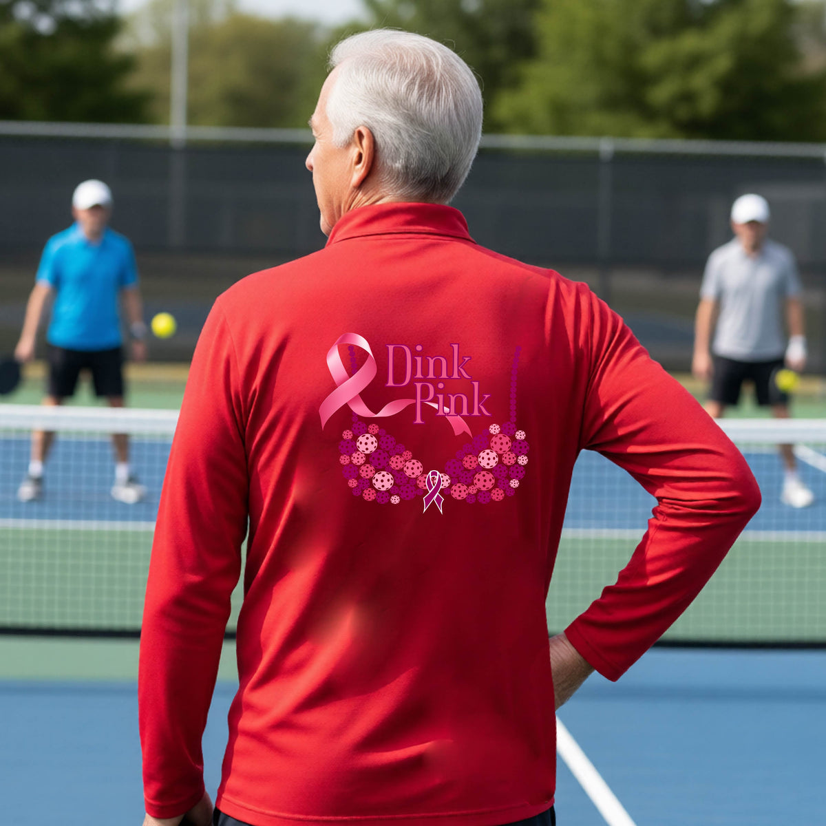 A man on a pickleball court seen from behind wearing a red long-sleeve quarter-zip shirt with a large pink 'Dink Pink' breast cancer awareness graphic made of pickleballs on the back.