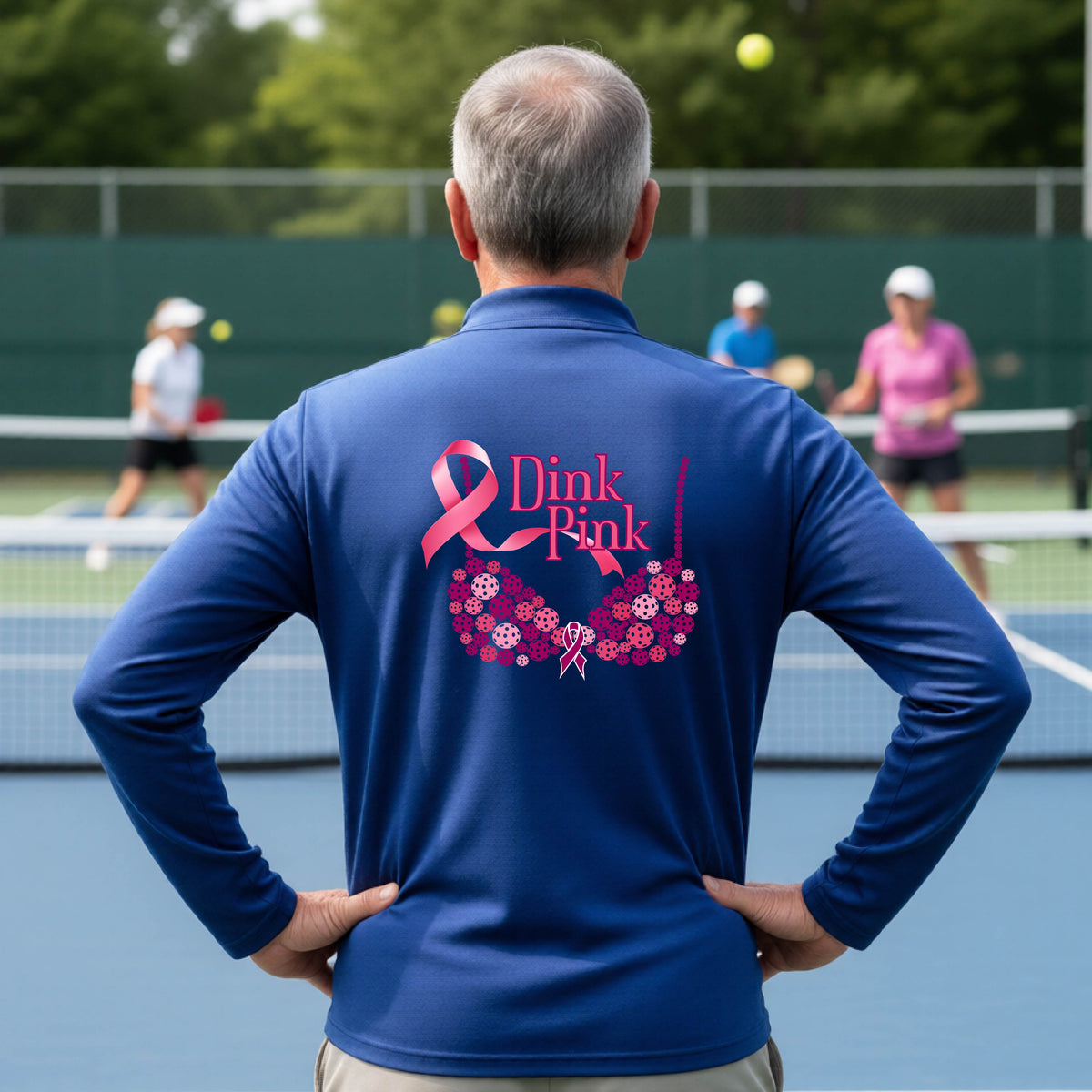 A man on a pickleball court seen from behind wearing a royal blue long-sleeve quarter-zip shirt with a large pink 'Dink Pink' breast cancer awareness graphic made of pickleballs on the back.