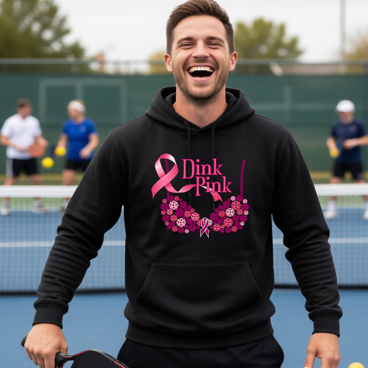 A man wearing a black hoodie with a pink 'Dink Pink' breast cancer awareness graphic made of pickleballs stands on a pickleball. The graphic features a pink ribbon, the words 'Dink Pink' in pink, and a floral bra design in shades of pink and purple.
