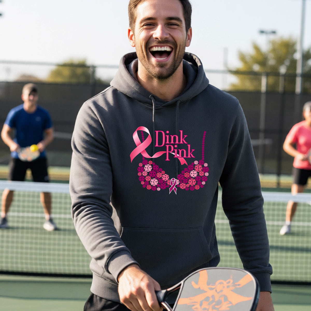 A man wearing a gray hoodie with a pink 'Dink Pink' breast cancer awareness graphic made of pickleballs stands on a pickleball. The graphic features a pink ribbon, the words 'Dink Pink' in pink, and a floral bra design in shades of pink and purple.