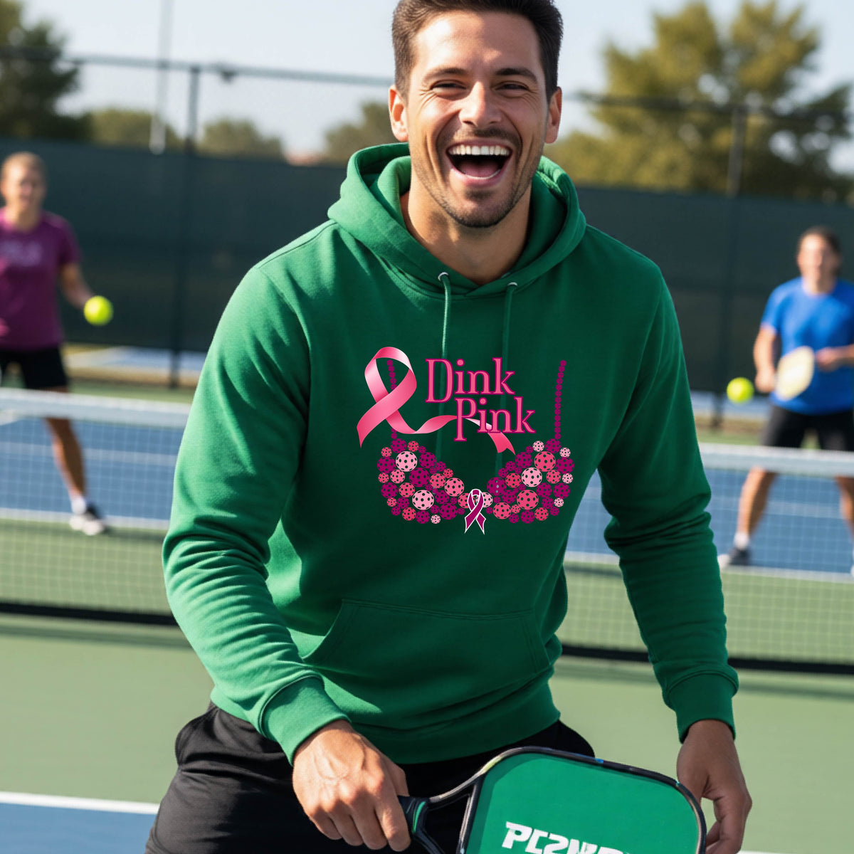 A man wearing a green hoodie with a pink 'Dink Pink' breast cancer awareness graphic made of pickleballs stands on a pickleball. The graphic features a pink ribbon, the words 'Dink Pink' in pink, and a floral bra design in shades of pink and purple.