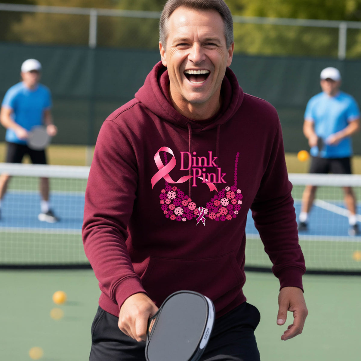 A man wearing a maroon hoodie with a pink 'Dink Pink' breast cancer awareness graphic made of pickleballs stands on a pickleball. The graphic features a pink ribbon, the words 'Dink Pink' in pink, and a floral bra design in shades of pink and purple.