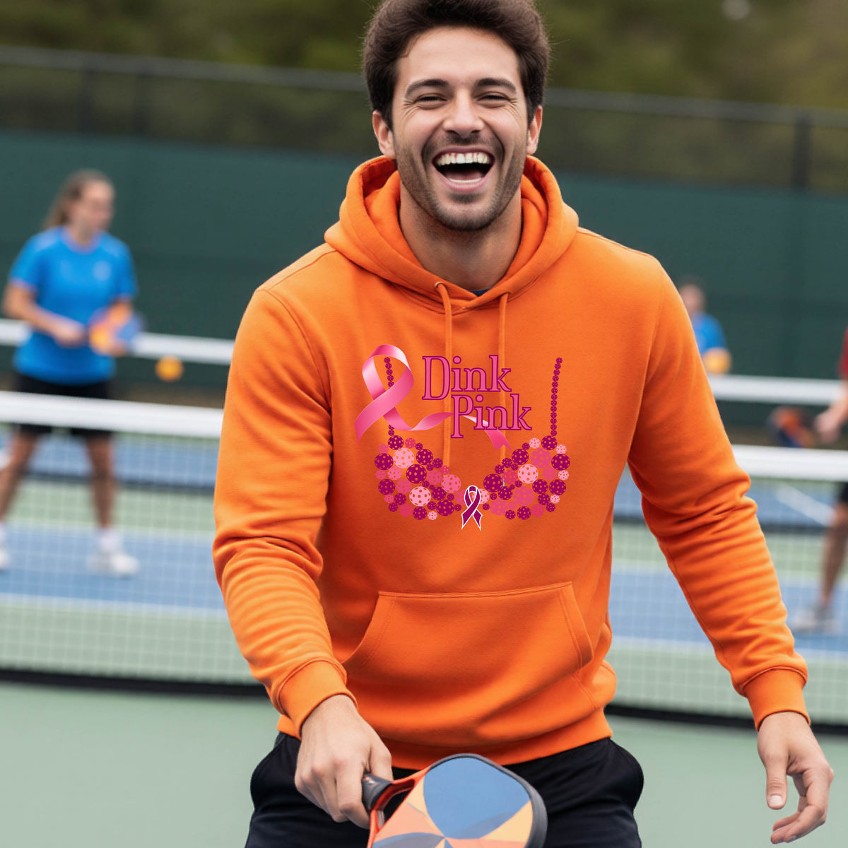 A man wearing an orange hoodie with a pink 'Dink Pink' breast cancer awareness graphic made of pickleballs stands on a pickleball. The graphic features a pink ribbon, the words 'Dink Pink' in pink, and a floral bra design in shades of pink and purple.