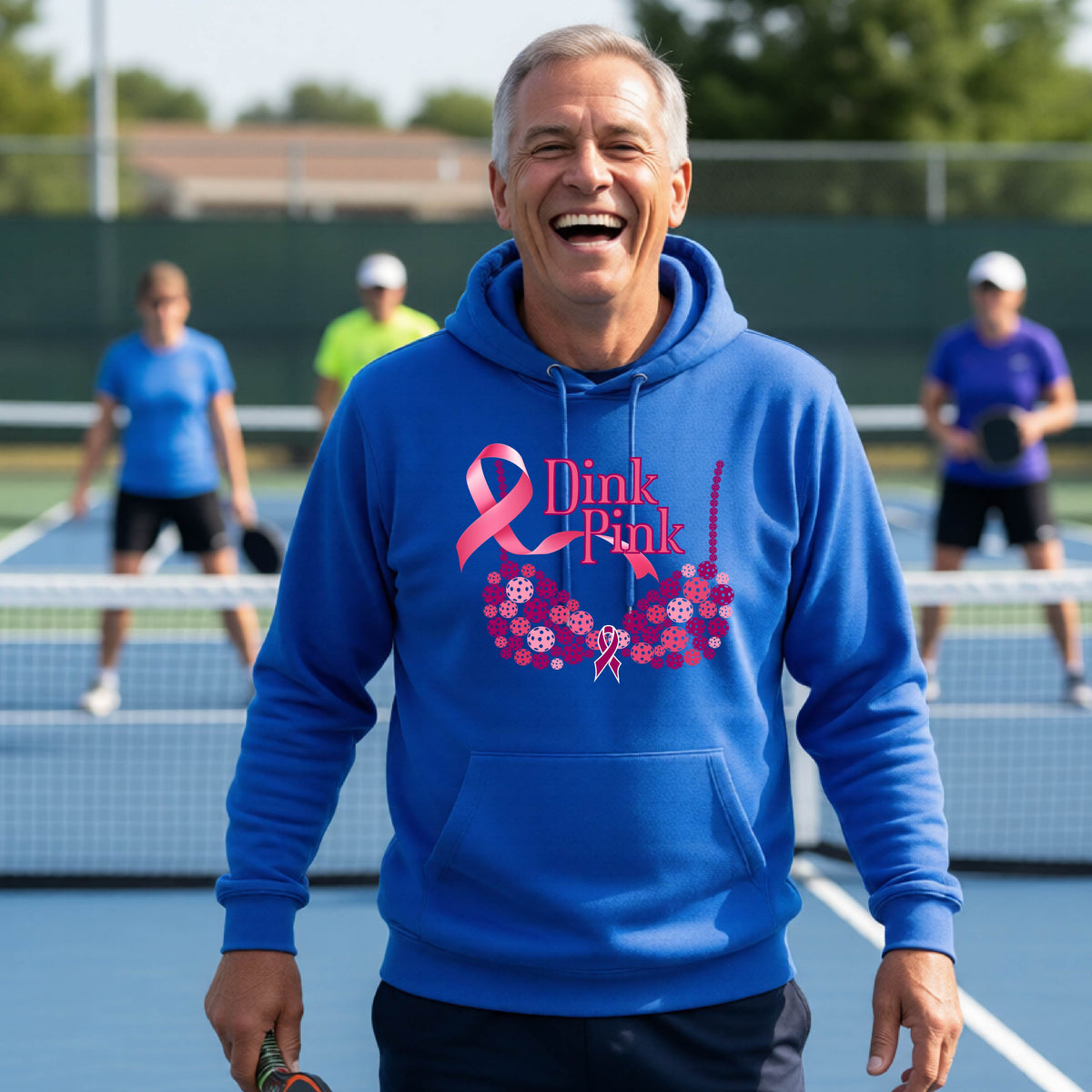 A man wearing a royal blue hoodie with a pink 'Dink Pink' breast cancer awareness graphic made of pickleballs stands on a pickleball. The graphic features a pink ribbon, the words 'Dink Pink' in pink, and a floral bra design in shades of pink and purple.