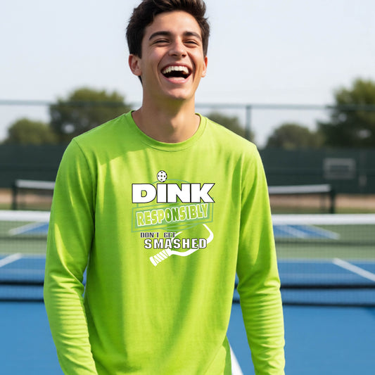 A young person laughing outdoor on a pickleball court, wearing a bright lime green long-sleeve shirt with a white and yellow graphic that reads 'DINK RESPONSIBLY DON'T GET SMASHED' with a whit pickleball paddle under the text, standing on a pickleball court. 
