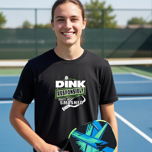 A young person wearing a black graphic t-shirt that reads 'DINK RESPONSIBLY DON'T GET SMASHED' in white and green with a white pickleball paddle under the text, standing on a pckleball court. 