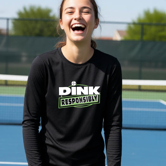 A young person laughing on an outdoor pickleball court wearing a black long-sleeve t-shirt with a white and green graphic that reads 'DINK RESPONSIBLY'.