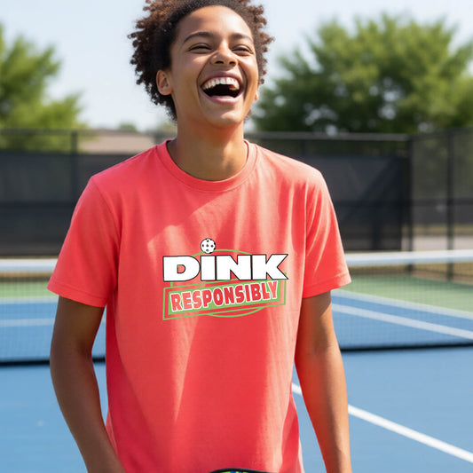 A smiling person standing on a pickleball court wearing a coral-orange graphic T-shirt with the words “DINK RESPONSIBLY” printed across the chest.