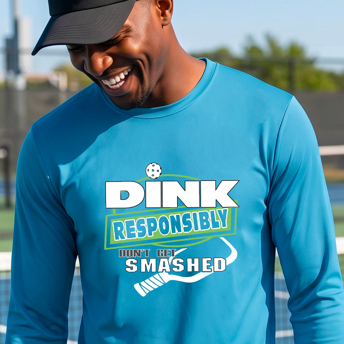 A man wearing a bright blue long-sleeve graphic t-shirt with a graphic of the text 'DINK RESPONSIBLY DON'T LET SMASHED' and a white pickleball paddle, standing on a pickleball court. 