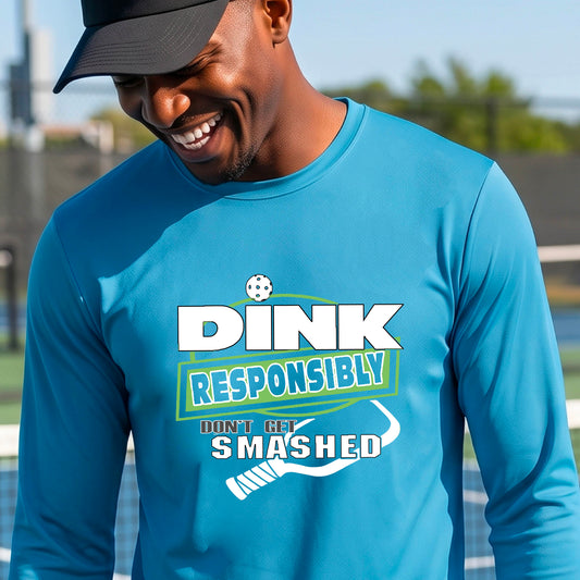 A man wearing a bright blue long-sleeve graphic t-shirt with a graphic of the text 'DINK RESPONSIBLY DON'T LET SMASHED' and a white pickleball paddle, standing on a pickleball court. 
