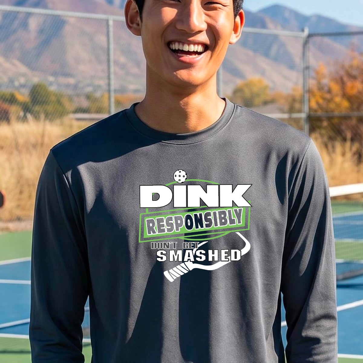 A man wearing a gray long-sleeve t-shirt with a 'DINK RESPONSIBLY DON'T LET SMASHED' graphic on the front, and a pickelball paddle graphic.