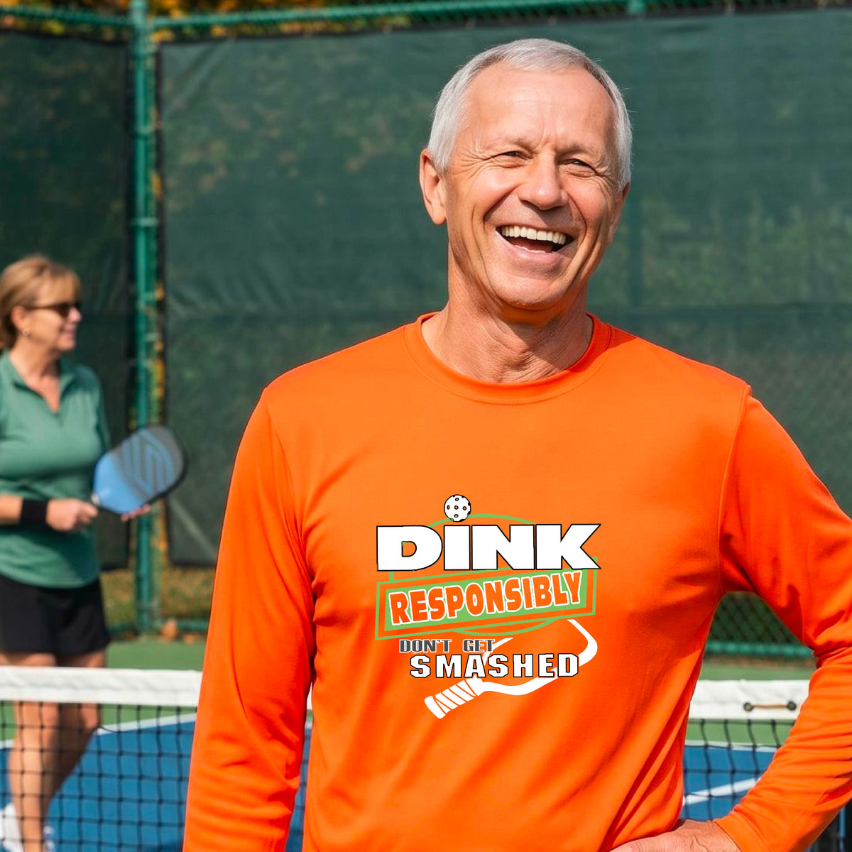 A man wearing a bright orange long-sleeve t-shirt with a 'DINK RESPONSIBLY DON'T LET SMASHED' graphic on the front, and a pickelball paddle graphic.