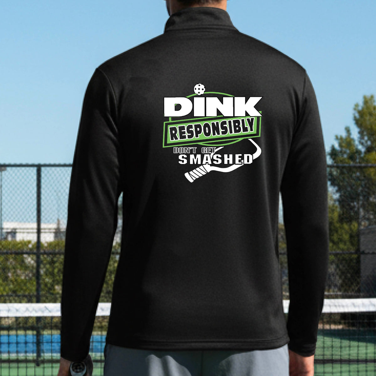 A man viewed from behind wearing a black long-sleeve athletic quarter-zip jacket with a large back graphic that reads 'DINK RESPONSIBLY DON'T LET SMASHED' with a white pickleball graphic on the design. 