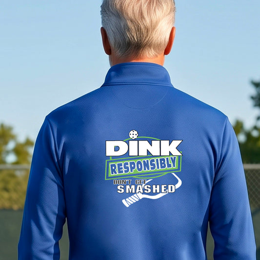 A man viewed from behind wearing a royal blue long-sleeve athletic quarter-zip jacket with a large back graphic that reads 'DINK RESPONSIBLY DON'T LET SMASHED' with a white pickleball graphic on the design. 