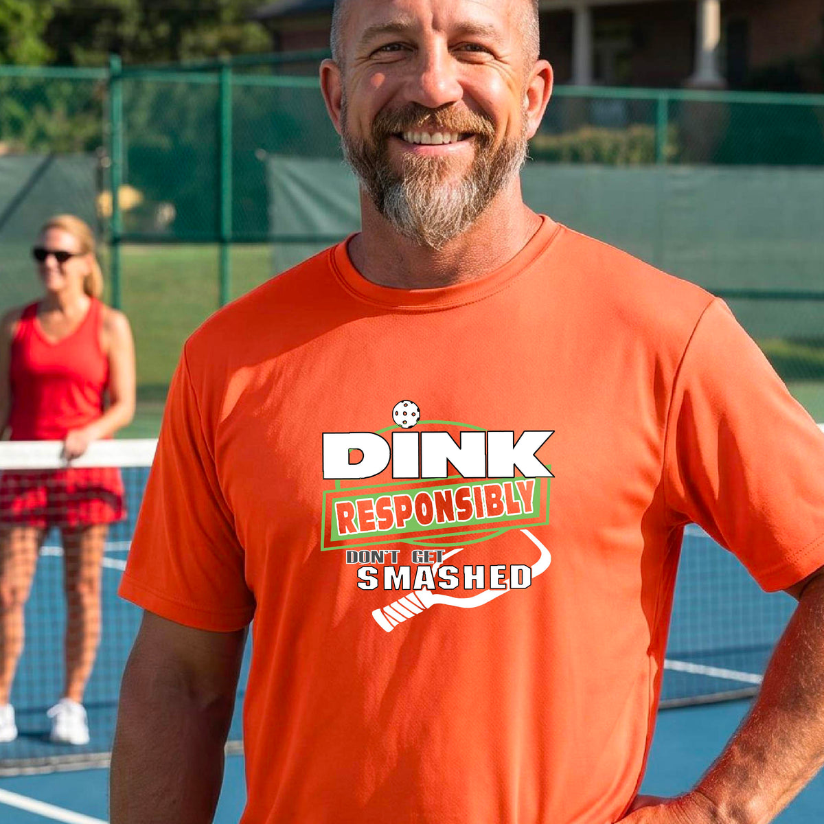 A smiling man wearing a bright orange graphic t-shirt with the words "DINK RESPONSIBLY DON'T GET SMASHED" with a white pickleball paddle under the text,standing on an outdoor pickleball court, 