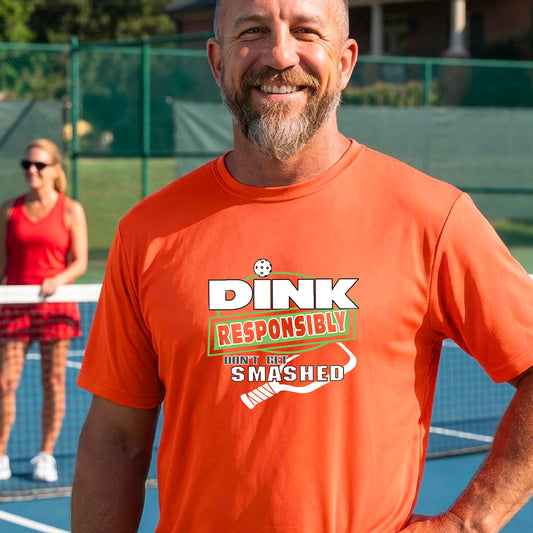 A smiling man wearing a bright orange graphic t-shirt with the words "DINK RESPONSIBLY DON'T GET SMASHED" with a white pickleball paddle under the text,standing on an outdoor pickleball court, 