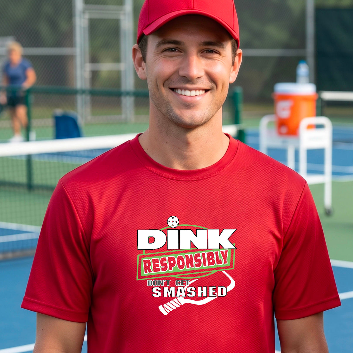 A smiling man wearing a red graphic t-shirt with the words "DINK RESPONSIBLY DON'T GET SMASHED" with a white pickleball paddle under the text,standing on an outdoor pickleball court, 