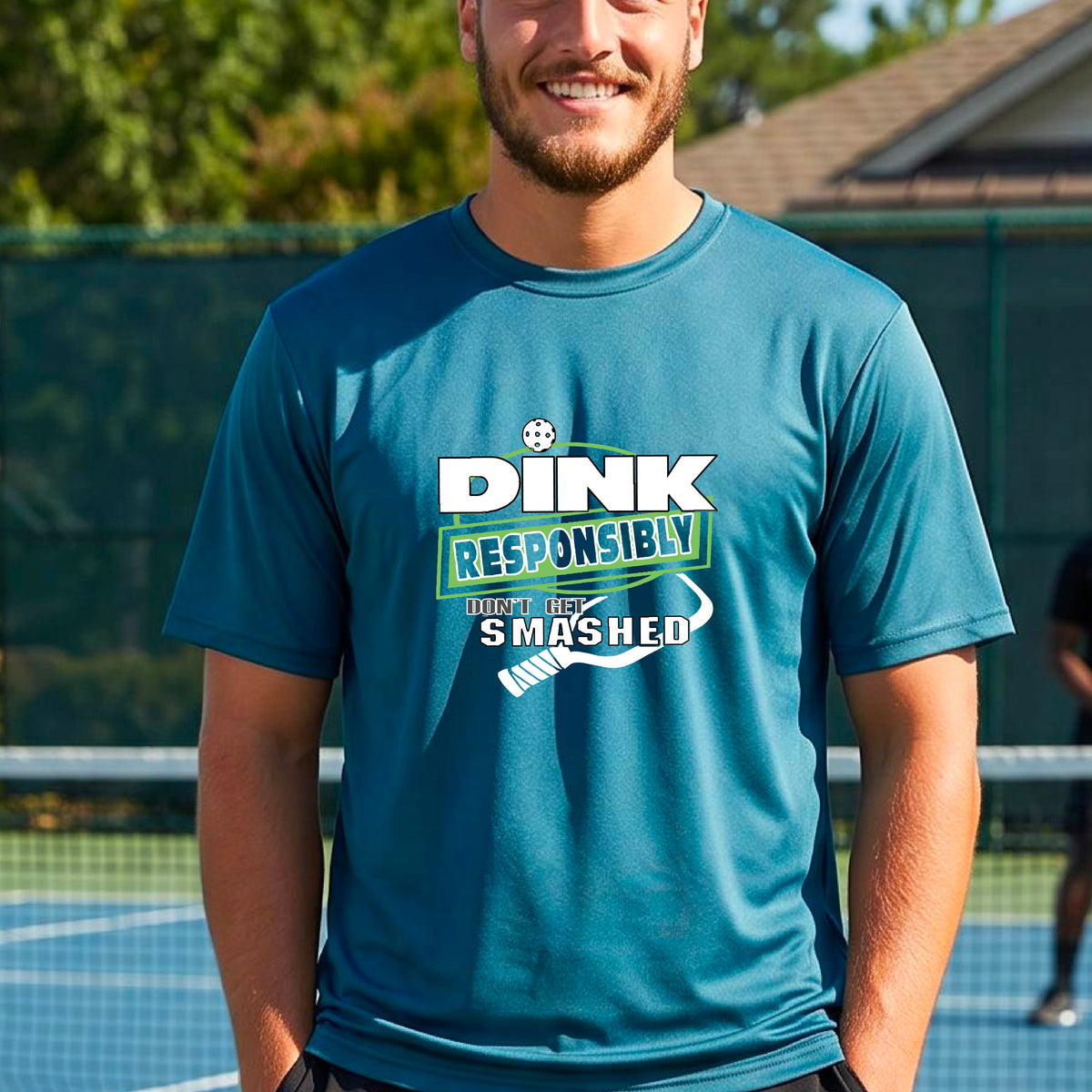 A smiling man wearing a teal graphic t-shirt with the words "DINK RESPONSIBLY DON'T GET SMASHED" with a white pickleball paddle under the text,standing on an outdoor pickleball court, 
