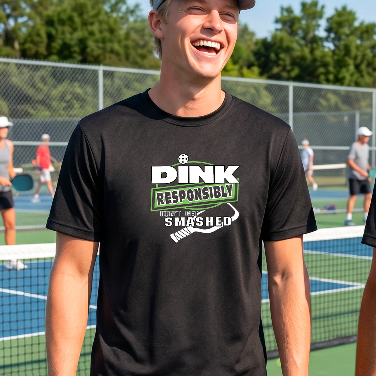 A smiling man wearing a black graphic t-shirt with the words "DINK RESPONSIBLY DON'T GET SMASHED" with a white pickleball paddle under the text,standing on an outdoor pickleball court, 